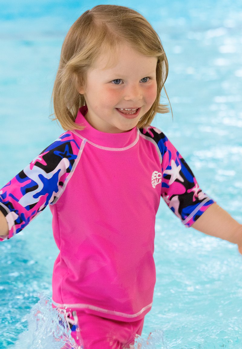 T-shirt de bain rose vif à manches courtes avec un motif de starfish colorés sur fond noir. Porté par un jeune enfant dans un cadre de piscine.