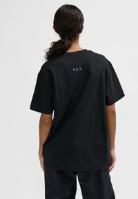 Schwarzes Baumwoll-T-Shirt mit kurzen Ärmeln, mit dem weißen Aufdruck "F.U.T." auf der Rückseite. Lockerer Schnitt und Rundhalsausschnitt.