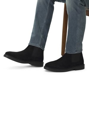 Schwarze Wildleder Chelsea-Stiefel mit elastischen Seitenpaneelen, flachen Sohlen und runder Zehe, getragen mit hellblauen Jeans, sitzend auf einem Holzstuhl.