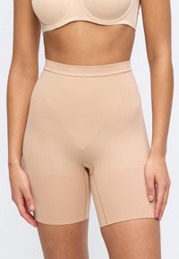 Γυναίκα που φοράει μπεζ σορτς υψηλής μέσης για σχηματισμό σώματος και κατάλληλο σουτιέν, στέκεται με τα χέρια χαλαρά στα πλάγια μπροστά σε απλό φόντο.