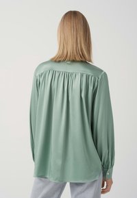 Camicia a maniche lunghe con colletto di colore verde giade, caratterizzata da un design con retro arricciato, texture liscia e polsini abbottonati.