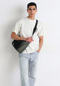 Sort og olivengrøn messenger taske med et slankt design, justerbar skulderrem og en frontklips, båret af en person i en lys t-shirt og jeans.