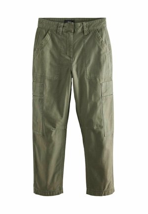 Femme debout à l'intérieur portant un top en maille gris sans manches, un pantalon cargo vert olive et des escarpins noirs à bout pointu, une main dans la poche.