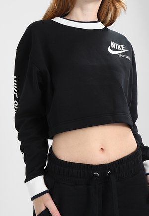 Kvinde iført sort kort Nike Sportswear sweatshirt med hvid krave og manchetter, kombineret med sorte bukser med snor, hånd i lommen.