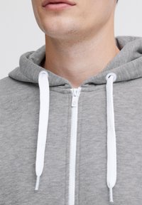 Felpa grigia con zip e bottone bianchi, realizzata in un tessuto morbido e strutturato. Presenta una zip frontale e un cappuccio. Vista ravvicinata del collo.