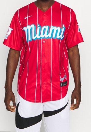 Homme portant un maillot de baseball rouge de Miami avec des rayures verticales blanches et un short blanc avec le logo noir de Nike.