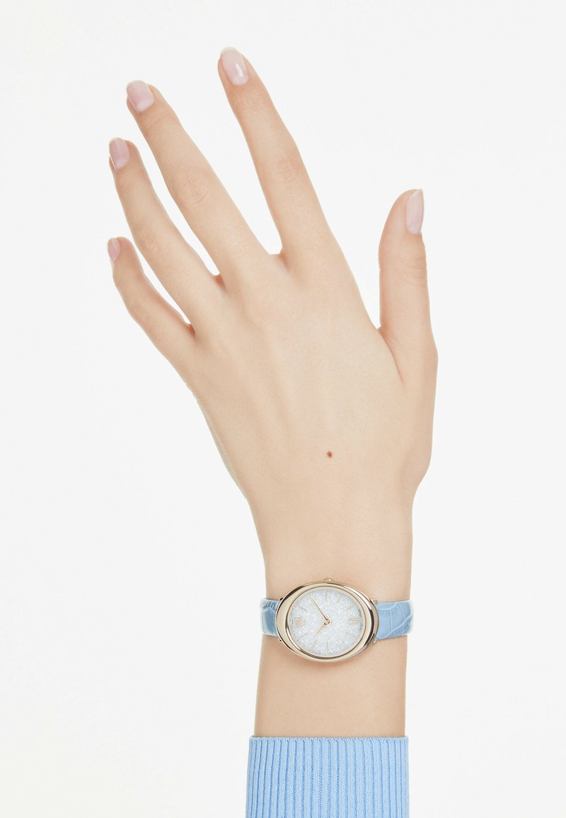 Orologio rotondo con quadrante argentato glitter, cassa dorata e cinturino in pelle stampata coccodrillo di colore azzurro chiaro. La mano è posizionata con le dita tese.