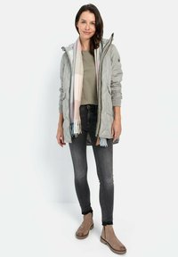 Graue gesteppte Jacke mit Kapuze, kombiniert mit einem beigen Oberteil, dunklen Jeans und hellbraunen Stiefeln. Der Schal hat ein pastellfarbenes Streifenmuster.