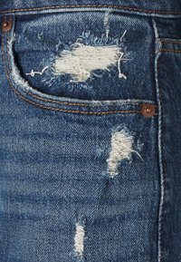 Denim-materiaali, jossa on keskisininen väri, ja johon on lisätty kuluneita alueita, purkautuvia reunoja ja paljastuneita valkoisia lankoja. Taskussa kuparinvärinen nappi.