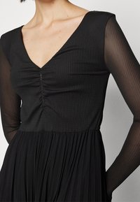 Žena v černých šatech s V-výstřihem a nařasením na hrudníku, se průhlednými dlouhými rukávy, stojící před jednobarevným pozadím.