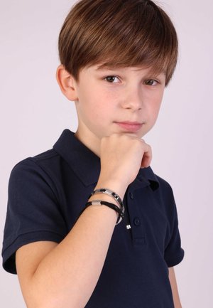 Zwarte en zilveren armband om een pols, met metalen schakels en een platte rechthoekige accent, gecombineerd met een marineblauwe poloshirt.