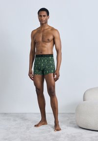 Zeleni bokser hlačke s črnim pasom. Tkanina ima gladko teksturo in vsebuje cikcak vzorec v različnih odtenkih zelene.