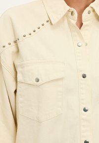 Camisa de mezclilla beige con tachuelas plateadas en los hombros, botones a presión y un bolsillo en el pecho con cierre de botón metálico.