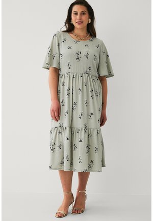 Kvinde iført mintgrøn midi-kjole med sort blomsterprint, korte flæseærmer og guld åbne sko, står mod en ensfarvet baggrund.