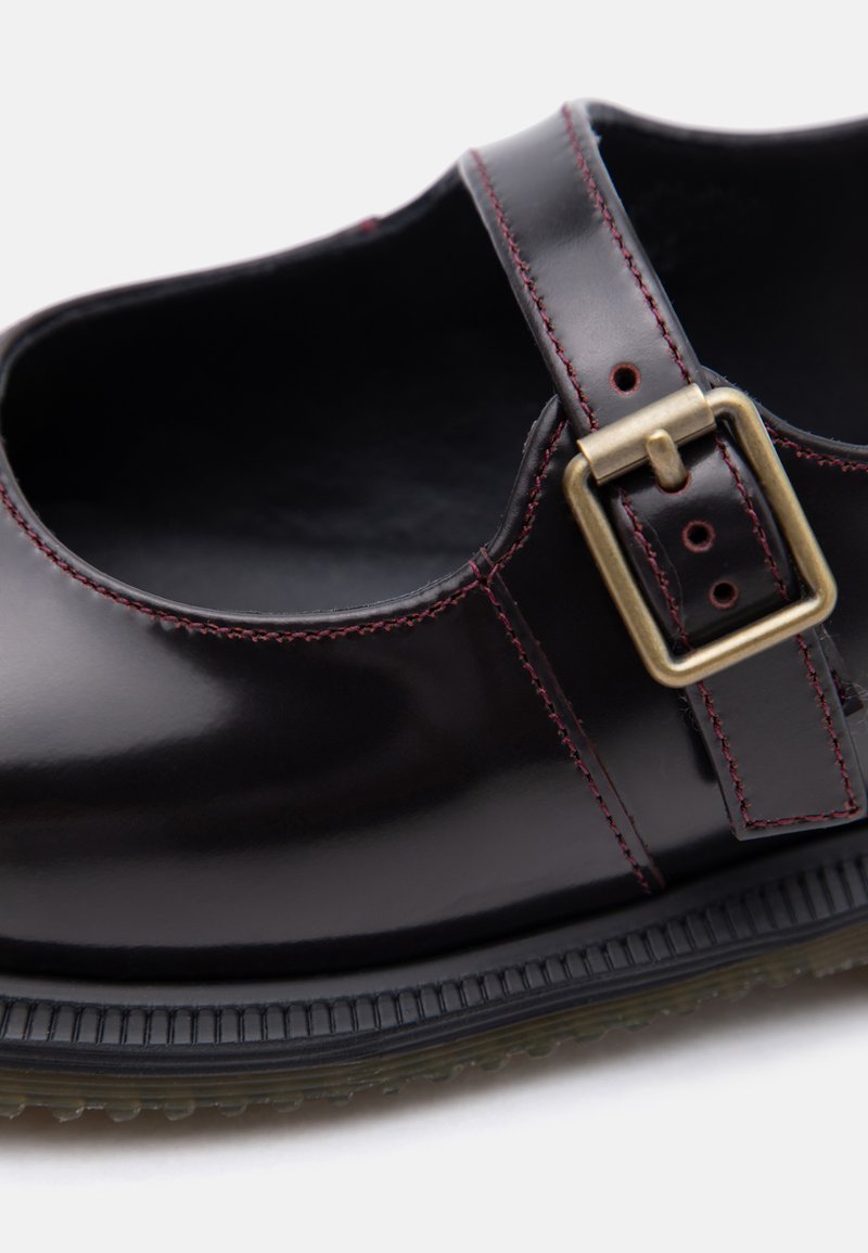Scarpa Mary Jane in pelle nera con fibbia in ottone, cuciture rosse e suola in gomma strutturata, mostrata in primo piano su sfondo bianco.