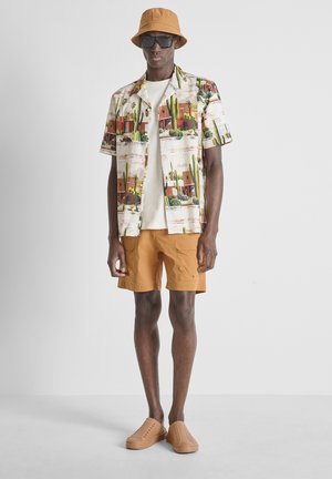 Homme portant un chapeau bob beige, des lunettes de soleil foncées, une chemise à manches courtes à motif cactus, un t-shirt blanc, un short beige et des chaussures beige sans lacets.