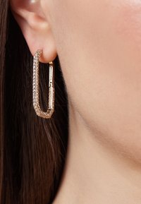 Boucle d'oreille géométrique en ton or, avec une surface texturée, ornée de strass transparents ; dotée d'un fermoir articulé pour un port sécurisé.