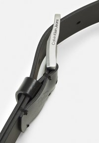 Cintura in pelle nera con fibbia in metallo argentato con incisione "Calvin Klein Jeans". Texture liscia con un design cucito.