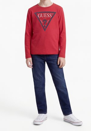 Ung person iført en rød Guess langærmet skjorte, blå jeans og hvide sneakers, stående mod en ensfarvet hvid baggrund.