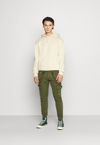 Sweat à capuche crème avec poche kangourou, associé à un pantalon cargo vert olive avec plusieurs poches et zips, complété par des baskets montantes vertes.