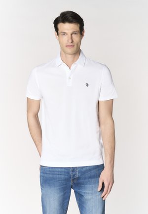 Uomo con capelli scuri che indossa una polo bianca a maniche corte con un piccolo logo e jeans blu, in piedi davanti a uno sfondo chiaro.