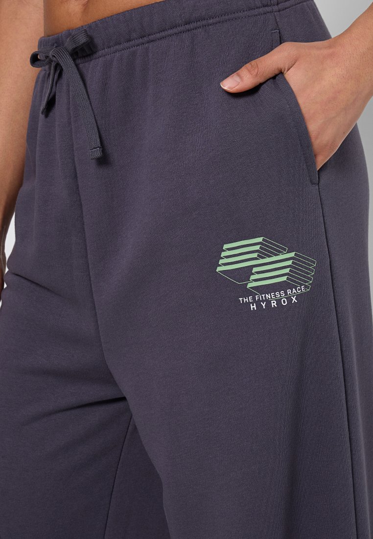 Dunkelgraue Sweatpants mit einem Kordelzugbund, die ein grünes Logo-Design auf dem linken Oberschenkel und seitliche Taschen besitzen. Weicher Stoff.