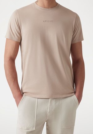 T-shirt beige à manches courtes avec logo « GUESS », porté avec un pantalon beige clair, mains dans les poches de manière décontractée, sur un fond uni.