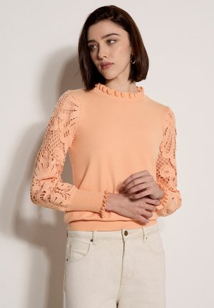 Femme aux cheveux bruns courts portant un pull pêche à manches en dentelle et un pantalon blanc, debout contre un mur clair uni.