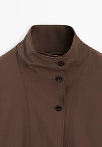 Brązowa kurtka z wysokim kołnierzem, zapinana na trzy czarne guziki z przodu, z widocznymi przeszyciami.