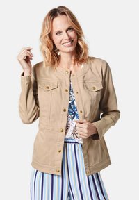 Beige jack met lange mouwen, drukknopen, twee borstzakken en een gladde textuur. Gecombineerd met gestreepte broek.