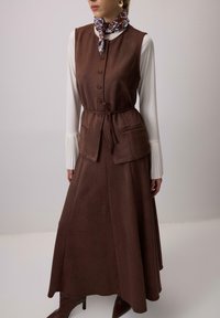 Rjava tkaninska telovnik z zavezovanjem v pasu, z gumbi spredaj in žepi, v kombinaciji z dolgo rjavo krilo. Bela bluza z nabranimi rokavi.