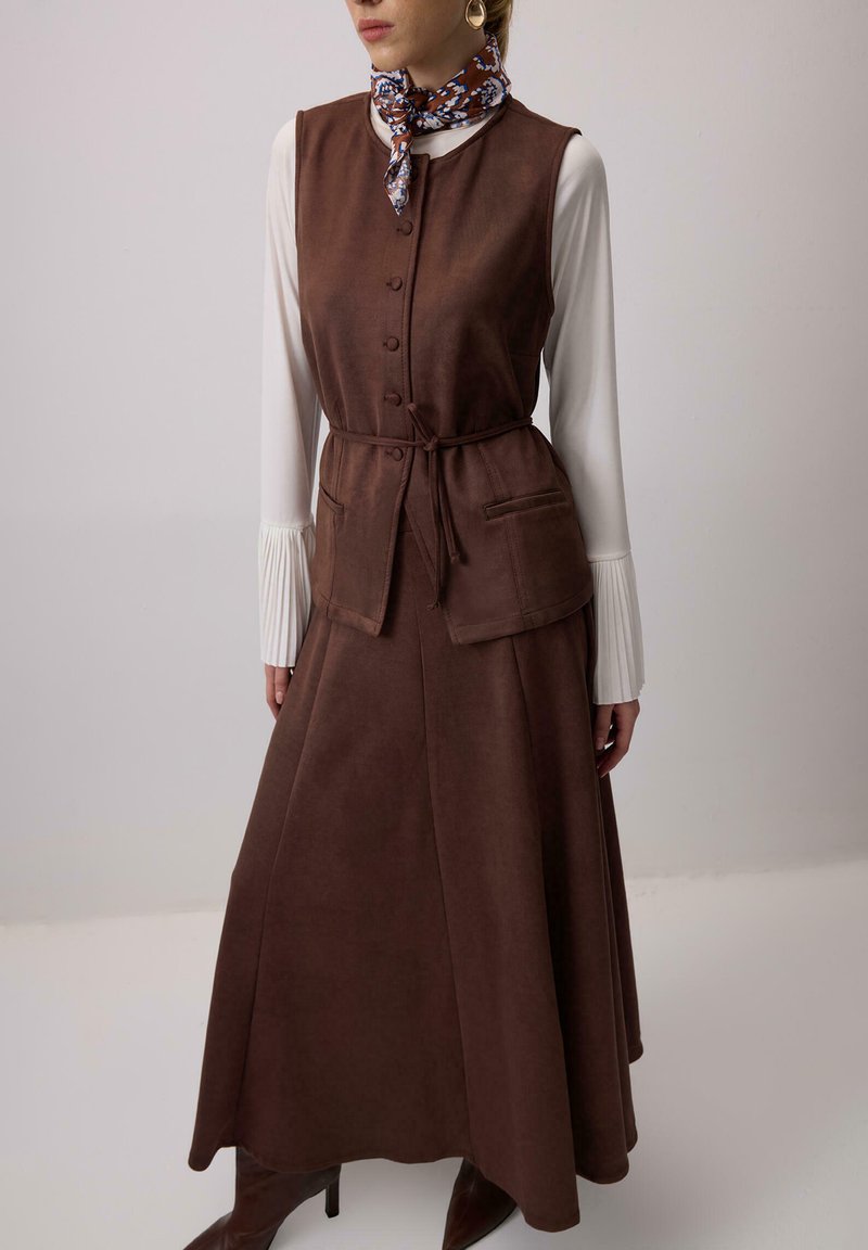 Rjava tkaninska telovnik z zavezovanjem v pasu, z gumbi spredaj in žepi, v kombinaciji z dolgo rjavo krilo. Bela bluza z nabranimi rokavi.