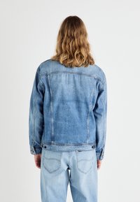 Ljusblå denimjacka med klassisk passform, synlig sömnad och knäppta manschetter. Strukturtyg visar lätt slitage, i kombination med ljusa jeans.