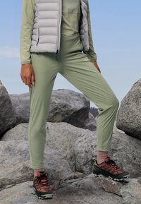 Completo sportivo verde chiaro con maglia a maniche lunghe e pantaloni a sigaretta, abbinato a un gilet isolante grigio e scarpe da trekking multicolore.
