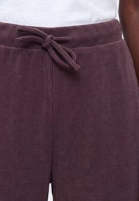 Gros plan d’un pantalon côtelé violet foncé avec une taille élastiquée nouée, porté avec un haut blanc.