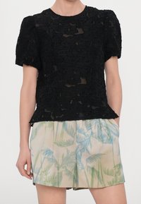 Top in tessuto nero con motivo floreale testurizzato, maniche puff corte e scollatura a girocollo, abbinato a pantaloni corti beige chiaro con stampa di foglie di palma.