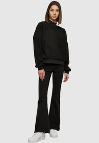Zwarte fleece pullover met een hoge halslijn, gecombineerd met zwarte flared jeans. Model draagt witte sneakers. Eenvoudig, comfortabel ontwerp.