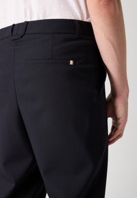 Dark blue trousers with a smooth texture, featuring a back pocket with a subtle striped accent in white and gold hues.