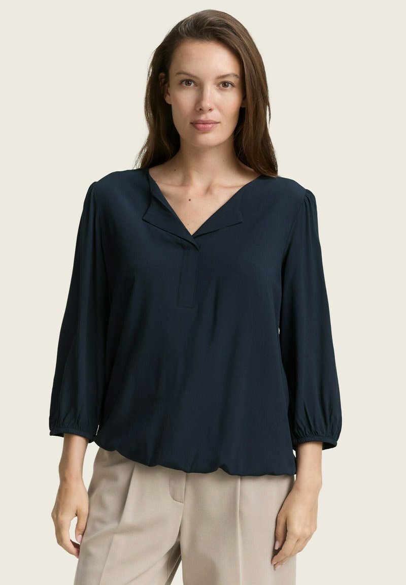 Blouse bleu marine avec un décolleté en V, des manches bouffantes trois-quarts, un tissu texturé et un ourlet froncé, associée à un pantalon beige clair.