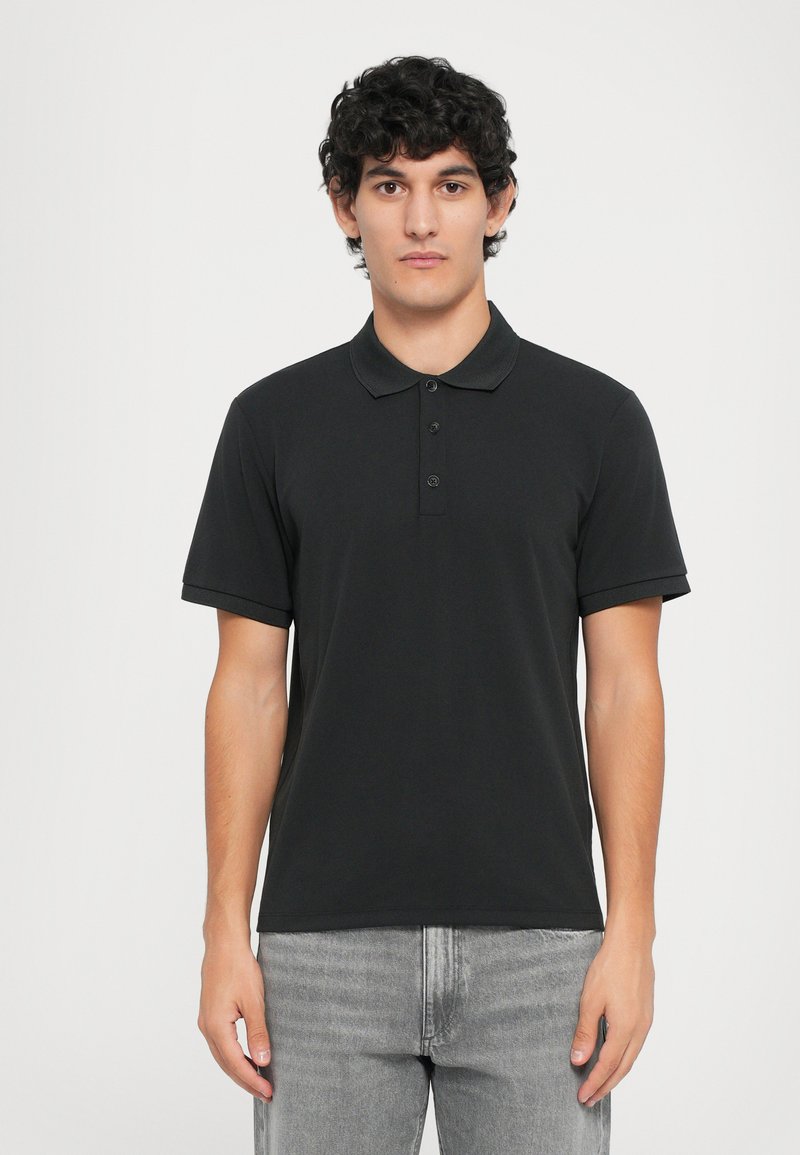 Polo noir avec un col, trois boutons à l'avant et des manches courtes. Porté avec un jean gris clair, mettant en avant un design simple.