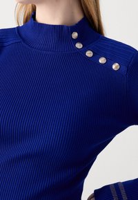 Gros plan sur une personne portant un pull côtelé bleu royal avec un col montant et cinq boutons décoratifs argentés sur l'épaule.