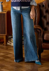 Platus kojų džinsai vidutinio mėlynumo džinso audinio, su aukštu liemeniu ir apsiūtais kraštais. Derinami su tamsiai mėlynu megztiniu su raštu ir žydrais batais.