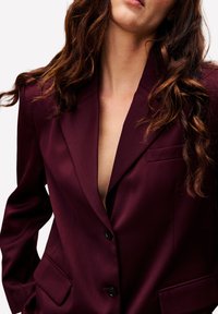 Vrouw met golvend bruin haar, gekleed in een diep bordeauxrode blazer met zwarte knopen, staande tegen een effen witte achtergrond.