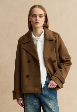 Giovane donna con capelli biondi lisci indossa una giacca doppiopetto marrone, una camicetta bianca con sciarpa e jeans blu con motivo floreale, in piedi davanti a uno sfondo beige.