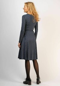 Marindress i marinblått med ett geometriskt mönster, långa ärmar, åtsittande midja, utställd kjol, bärs med svarta strumpbyxor och kraftiga klackar. Slät textur.