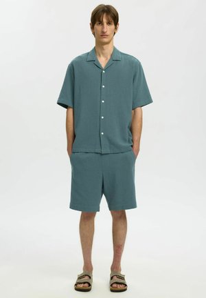 Młody mężczyzna stoi z rękami w kieszeniach, ubrany w turkusową koszulę na krótki rękaw z guzikami, dopasowane szorty i beżowe sandały na jednolitym tle.