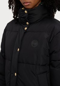 Veste rembourrée noire avec un col haut, des boutons pression dorés et un écusson rond sur la poitrine. Texture lisse et design matelassé.