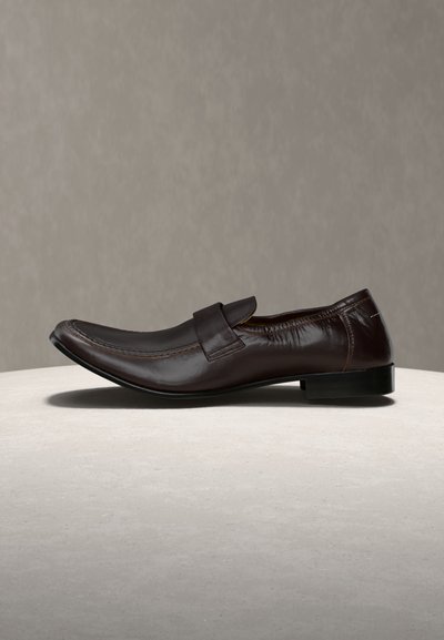 Καφέ σκούρο δερμάτινο ανδρικό μοκασίνι με κομψό σχεδιασμό και χαμηλό τακούνι, τοποθετημένο σε ανοιχτόχρωμη στρογγυλή επιφάνεια πάνω σε ουδέτερο φόντο.