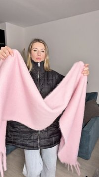 Sciarpa rosa, morbida, con frange, tenuta a forma di V, realizzata in un materiale soffice. Giacca nera trapuntata indossata sotto, con chiusura a zip.