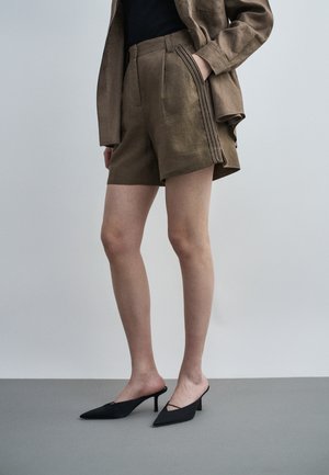 Osoba nosiaca hnedé textúrované šortky s dekoratívnymi pruhmi na boku, zodpovedajúcu bundu, čierny top a čierne nízke lodičky na podpätku so špicatou špičkou.